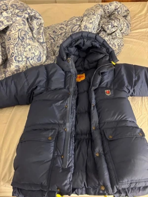 Mörkblå dunjacka från Fjällräven - Varm mörkblå dunjacka från Fjällräven med stor huva, två stora framfickor med knappar och dragkedja. Jackan har klassisk Fjällräven-logga på bröstet och gula detaljer vid dragkedjan. Perfekt för kalla vinterdagar. En tryckknapp är trasig på insidan av en ficka, inget som syns utåt. I övrigt väldigt lite använd. 