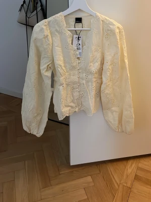 Gul blus med broderade detaljer XS - Söt gul blus med ballongärmar och broderade blommor. Blusen har knappar framtill och en feminin, figurnära passform. Perfekt för dig som gillar romantisk och trendig stil. Materialet känns lätt och luftigt.