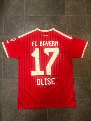 Bayern München OLISE #17 Adidas Fotbollströja - Säljer en röd Bayern München matchtröja från Adidas med vita detaljer och klubbmärke på bröstet. Michael Olise på ryggen #17. Tröjan har korta ärmar, rund halsringning med guldkant och vita ränder på axlarna. Tillverkad i lätt och ventilerande material, perfekt för fotbollsträning, match eller bara en helt vanlig utekväll. 