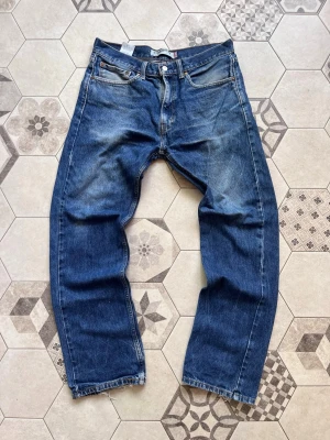Levi's 501 blå raka jeans - Klassiska blå Levi's 501 jeans med rak passform och knappgylf. Jeansen har fem fickor, tydliga slitningar och den ikoniska läderlappen bak i midjan. Tillverkade i kraftig denim med vintagekänsla och snygg tvätt.