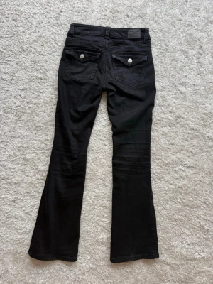 Svarta bootcut jeans med fickdetaljer - Säljer ett par svarta bootcut jeans med snygga fickor bak som har lock och silverknappar. Jeansen har klassisk femficksdesign och är i stretchigt material för skön passform. Perfekta till en trendig och avslappnad look.