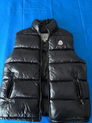 Svart dunväst från Moncler - Snygg svart dunväst från Moncler med glansig finish och klassisk puffer-design. Västen har två dragkedjefickor framtill och hög krage. Moncler-logga på bröstet ger en exklusiv touch. Perfekt att slänga över en hoodie eller tröja.