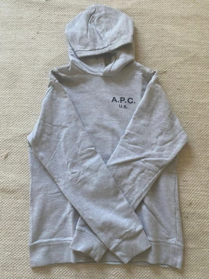 Grå hoodie från A.P.C. med tryck - Snygg ljusgrå hoodie från A.P.C. med svart tryck 'A.P.C. U.S.' på bröstet. Klassisk modell med huva och ribbade muddar vid ärmslut och nederkant. Perfekt för en avslappnad och stilren look. Materialet känns mjukt och bekvämt.