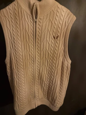 Beige kabelstickad väst med dragkedja - Säljer en beige kabelstickad väst med hög krage och dragkedja framtill. Västen har ett diskret broderat emblem på bröstet och är tillverkad i ett mjukt, tjockt material. Perfekt för lager på lager och ger en klassisk look.
