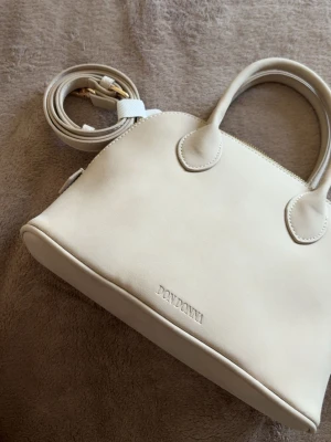 Beige handväska Don Donna - Snygg beige handväska från Don Donna i mjukt material med rundad form. Väskan har två handtag och avtagbar axelrem i matchande färg. Köpte den för 499kr, kvitto finns kvar. Aldrig använd