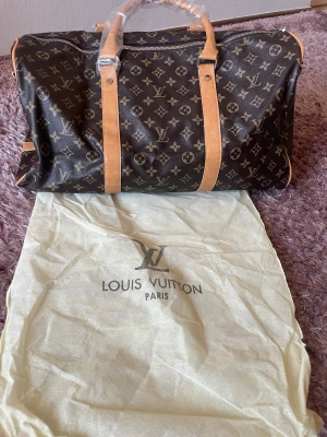 Resväska - Säljer en klassisk resväska i brunt med ikoniskt monogrammönster och ljusbruna läderdetaljer. Väskan har dubbla handtag, dragkedja och rymligt fack med innerficka. Perfekt för weekendresor och stilrena äventyr.