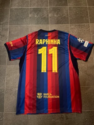 Barcelona matchtröja Raphinha #11 - Säljer en FC Barcelona matchtröja från Nike med Raphinha och nummer 11 på ryggen. Tröjan har klassiska blå och röda vertikala ränder, Champions League-märke på ärmen och Spotify-logga på bröstet. Tillverkad i lätt och ventilerande material. På tröjan står det XL men den sitter defenetivt mer som en L.