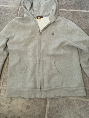 Grå hoodie från Polo Ralph Lauren - Klassisk grå hoodie från Polo Ralph Lauren med dragkedja framtill och huva. Har två fickor och den ikoniska lilla loggan broderad på bröstet. Perfekt för en chill och avslappnad stil.