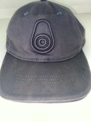 Mörkgrå keps med broderad logga - Snygg mörkgrå keps i bomull med unik broderad logga framtill. Klassisk böjd skärm och justerbar rem bak för perfekt passform. Ventilationshål upptill för extra komfort. Passar dig som gillar stilrena accessoarer.