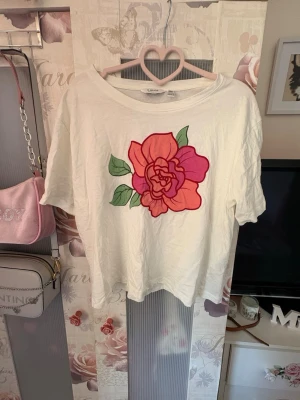 Vit t-shirt med stor blomma - Säljer en vit t-shirt med en stor, färgglad blomma i rosa och rött på bröstet. T-shirten har korta ärmar och en avslappnad passform. Perfekt för dig som gillar statement-plagg och vill ha något som sticker ut i garderoben.