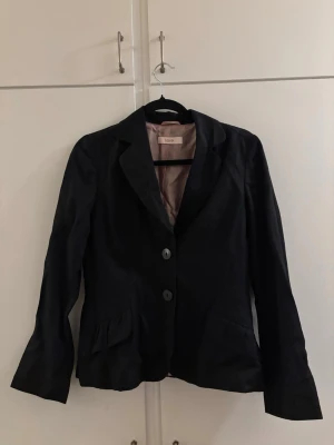 Svart kavaj från Laurèl - Stilren svart kavaj från Laurèl med två knappar framtill och klassisk krage. Kavajen har två fickor med diskreta veckdetaljer och ett ljusrosa foder. Perfekt för en dressad look och enkel att matcha med olika outfits.