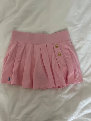 Rosa plisserad kjol från Polo Ralph Lauren - Supersöt rosa kjol från Polo Ralph Lauren med bred midjeresår, två guldknappar framtill och plisserad design. Kjolen har en liten broderad logga i blått nedtill och är gjord i mjuk bomull som känns skön mot huden. Kjolen har inbyggda shorts 