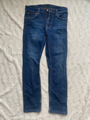 Mörkblå skinny jeans från Nudie Jeans - Snygga mörkblå skinny jeans från Nudie Jeans med klassisk femficksmodell och kontrastsömmar. Jeansen har en smal passform och är tillverkade i mjukt denimtyg. Perfekta för dig som gillar en stilren och tidlös look. W27 L 30