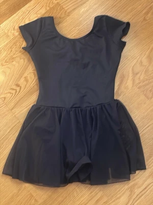 Svart balettklänning från Capezio för barn - Säljer en svart balettklänning från Capezio i storlek M. Klänningen har korta ärmar, rund halsringning och en kjoldel i tunt, genomskinligt tyg. Perfekt för dans eller träning. Materialet är stretchigt och följsamt. För barn