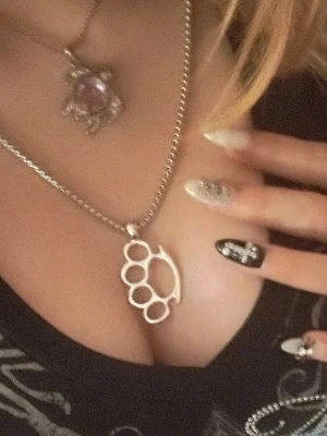 Silverfärgat halsband med knogjärn - Snyggt halsband i silverfärg med ett hänge format som ett knogjärn. Kedjan är tunn och stilren, perfekt för dig som gillar edgy accessoarer. Passar till många olika stilar och ger en cool touch till din outfit.