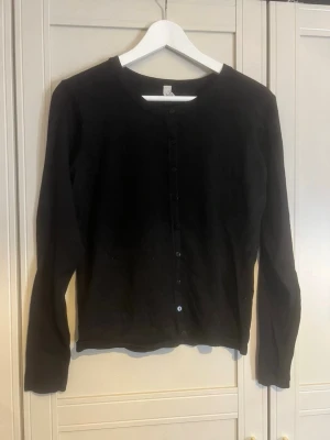 Svart basic kofta med knappar - En enkel svart kofta med rund halsringning och knäppning framtill. Långärmad modell i mjukt material, perfekt att slänga över en t-shirt eller linne. Stilren och lättmatchad till det mesta i garderoben. Har katt.