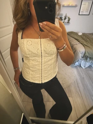 Vit broderad bustier topp - Söt vit bustier topp med breda axelband och broderade blommönster. Toppen har en rak skärning vid bysten och dragkedja framtill. Tillverkad i ett lätt bomullsliknande material som känns luftigt och somrigt.