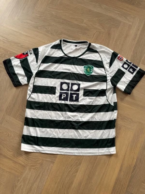 Sporting Lissabon fotbollströja Ronaldo - Grönvit randig fotbollströja från Sporting Lissabon med klubbmärke på bröstet och sponsorloggor. På ryggen står det 'C. Ronaldo' och nummer 28. Tröjan har korta ärmar och rund hals, tillverkad i lätt och ventilerande material. Stolen XL men sitter som L