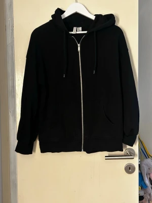 Svart zip hoodie med huva - En klassisk svart hoodie med dragkedja framtill och huva med snören. Hoodien har två fickor på magen och ribbade muddar vid ärmar och nederkant. Perfekt för en avslappnad och stilren look. Storlek xs oversized så passar även större storlekar