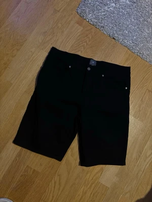 Svarta slim fit shorts från H&M - Säljer ett par svarta slim fit shorts från H&M med klassisk femficksdesign och knappgylf. De har en enkel och clean look som passar till det mesta. Materialet är mjukt och stretchigt för extra komfort.