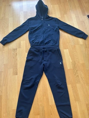 Polo Ralph Lauren tracksuit / mjukisset marinblå  - Säljer ett stilrent marinblått mjukisset från Polo Ralph Lauren bestående av zip-hoodie och matchande byxor.  Båda delarna är storlek S och har den klassiska broderade Polo-loggan. Hoodien har hel dragkedja, luva och fickor. Byxorna har dragsko i midjan och sidofickor.  Setet är begagnat men i fint skick. Se bilder för skick. Kvitto finns. 