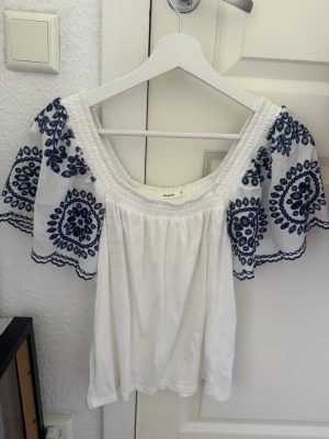 Vit blus med blå broderi från Reserved - Supersöt vit blus från Reserved med vida korta ärmar och blå broderade mönster. Blusen har en bred, smockad urringning och är perfekt för sommarens alla tillfällen. Luftigt material och snygga detaljer på ärmarna.