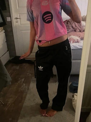 Svarta Adidas mjukisbyxor med vita ränder - Säljer ett par svarta Adidas mjukisbyxor med klassiska vita ränder längs sidorna och Adidas-logga på vänster ben. Byxorna har avslappnad passform och resår i midjan. Perfekta för chill dagar eller träning. Oklart vilken storlek men passar S!