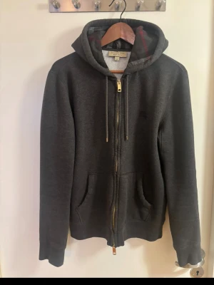 Burberry Hoodie - Säljer en mörkgrå zip-hoodie från Burberry i storlek M.  Klassisk modell med Burberrys rutiga mönster på insidan av huvan, diskret broderad Burberry-logga på bröstet samt guldfärgade detaljer på dragkedja och snören.  Hoodien är begagnad men i fint skick. Se bilder för skick.  Storlek: M Färg: mörkgrå Original Burberry Köpt på NK Kvitto finns och kan visas vid seriöst intresse.