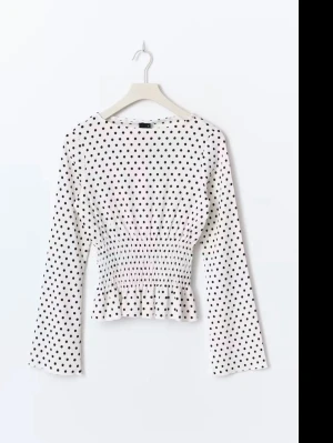 Gina Tricot blus  - Söt vit blus med svarta prickar och smockad midja som ger en snygg siluett. Blusen har långa ärmar och en liten volang längst ner. Perfekt för dig som gillar trendiga detaljer och klassiskt mönster.