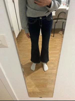 Mörkblå bootcut jeans med kontrastsömmar - Säljer ett par mörkblå bootcut jeans med vita kontrastsömmar och snygga fickdetaljer bak. Jeansen har låg midja och vida ben, vilket ger en retro och trendig look. Perfekta för dig som vill sticka ut med en unik jeansstil.
