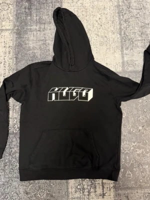 Svart hoodie med HUGO-tryck - Snygg svart hoodie från HUGO med stort vitt 3D-tryck på bröstet. Klassisk känguruficka framtill och mjuk huva. Perfekt för dig som gillar streetwear och vill ha en stilren men ändå iögonfallande look.