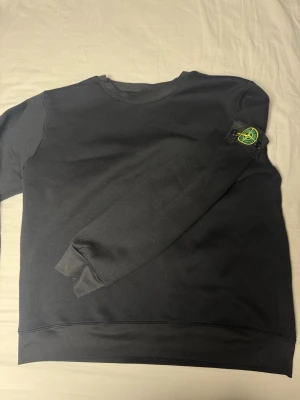Svart sweatshirt från Stone Island - Säljer en svart sweatshirt från Stone Island med den klassiska loggan på vänster ärm. Tröjan har rund hals, ribbade muddar och är tillverkad i mjukt bomullsmaterial. Enkel och stilren design som funkar till det mesta.
