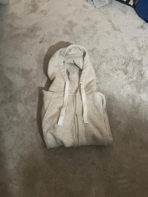 Ljusgrå zip hoodie med fickor - En stilren ljusgrå hoodie med dragkedja framtill och två stora fickor. Tröjan har huva med vita snören och ribbade muddar vid ärmar och nederkant. Perfekt för en avslappnad look och enkel att matcha med det mesta.
