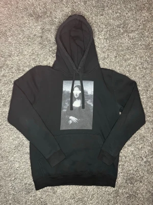 Svart hoodie med Mona Lisa-tryck - Svart hoodie med stor huva och snörning. På bröstet finns ett stort tryck av Mona Lisa i svartvitt. Klassisk känguruficka framtill och ribbade muddar vid ärmar och nederkant. Perfekt för dig som gillar streetwear och konstinspirerade plagg.