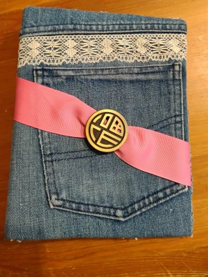 Handgjord jeansklädd pärm - Unik handgjord pärm klädd i blått jeanstyg med spetsdetalj upptill och en funktionell ficka framtill. Dekorerad med ett brett rosa band och en stor dekorativ knapp. Perfekt för anteckningar eller som personlig kalender. A5 storlek