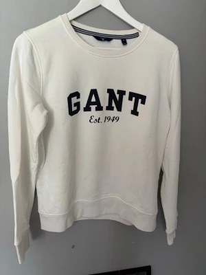 Vit sweatshirt från GANT - Klassisk vit sweatshirt från GANT med svart broderad logga och texten 'Est. 1949' på bröstet. Tröjan har rund halsringning, ribbade muddar och är tillverkad i mjukt bomullsmaterial. Perfekt för en clean och stilren look.