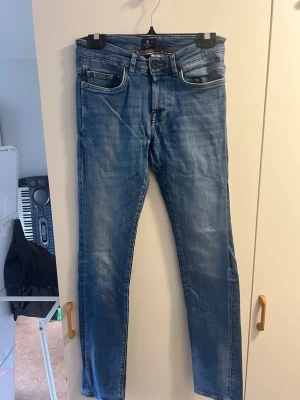 Ljusblå raka jeans med fem fickor - Säljer ett par ljusblå jeans med klassisk femficksmodell och raka ben. Jeansen har snygga slitningar och kontrastsömmar, samt en knappgylf. Tillverkade i denim, perfekt för dig som gillar en avslappnad och tidlös stil.