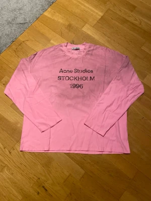 Rosa Acne Studios sleeve - Säljer en rosa långärmad t-shirt från Acne Studios med tryck 'STOCKHOLM 1996' på bröstet. Klassisk rund hals och relaxed fit. Perfekt för dig som gillar streetwear och vill ha något unikt från ett svenskt märke.