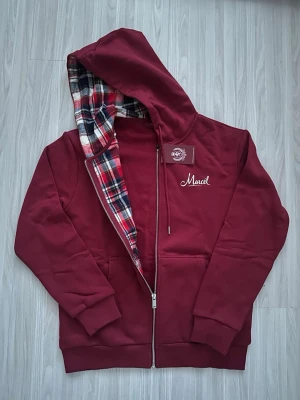 Vinröd hoodie med rutigt foder - Snygg vinröd hoodie med dragkedja och huva. Insidan av huvan har ett rutigt foder i rött, vitt och svart. På bröstet finns en liten vit brodyr med texten 'Marcel'. Hoodien har fickor framtill och ribbade muddar.