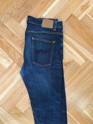 Mörkblå smala jeans från Nudie - Snygga mörkblå jeans från Nudie med klassisk femficksdesign och kontrastsömmar. Jeansen har smal passform och är tillverkade i stretchigt denimtyg för extra komfort. Bakfickorna har Nudies ikoniska vågmönster. Perfekta till din vardagsstil. (Dem har blivit lagade se bild 4)