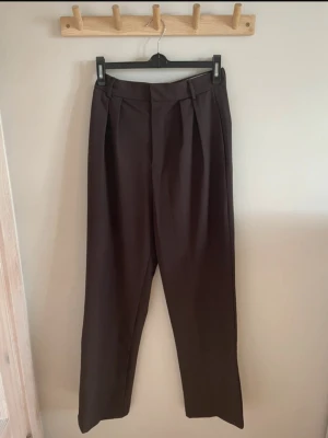 Bruna kostymbyxor med pressveck - Snygga bruna kostymbyxor med raka ben och pressveck framtill. Byxorna har hög midja, bälteshällor och veckade detaljer vid linningen för en stilren look. Perfekta för dig som gillar klassisk men trendig stil.