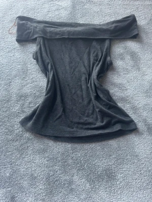 Mörkgrå offshoulder topp Kappahl - Säljer en mörkgrå offshoulder topp från Kappahl i mjukt material. Toppen har en axelbandslös design och sitter snyggt över axlarna. Perfekt för dig som gillar en enkel men trendig stil.