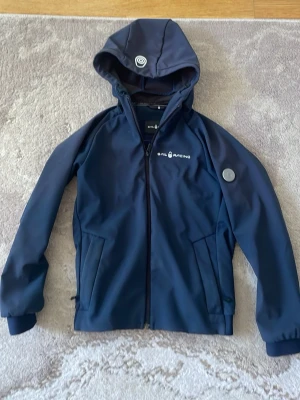 Sail Racing höst jacka - Snygg marinblå höst jacka   från Sail Racing med luva  och dragkedja framtill. Jackan har två sidofickor med dragkedja och logga på bröstet samt en detalj på armen. Materialet är slätt och vattenavvisande, perfekt för blåsiga dagar.