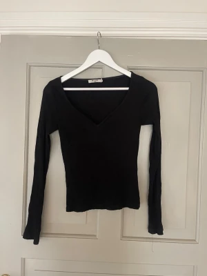Svart ribbad topp från NA-KD - Snygg svart långärmad topp från NA-KD med djup v-ringning och ribbad struktur. Perfekt att styla med jeans eller kjol för en trendig look. Tillverkad i mjukt stretchigt material som sitter väldigt snyggt!
