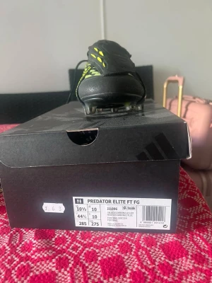 Adidas Predator Elite FT FG fotbollsskor - Säljer ett par Adidas Predator Elite FT FG fotbollsskor i svart med gula och vita detaljer. Skorna har låg profil, snörning och mönstrad ovandel för extra grepp. Perfekta för spel på fasta underlag. Snygg och modern design för dig som vill sticka ut på planen.