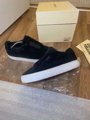 Svarta sneakers från Axel Arigato - Stilrena svarta sneakers från Axel Arigato i mocka med vit sula och diskret logga på sidan. Klassisk låg modell med minimalistisk design och elastiska band istället för vanliga skosnören. Perfekta för en clean och modern look.