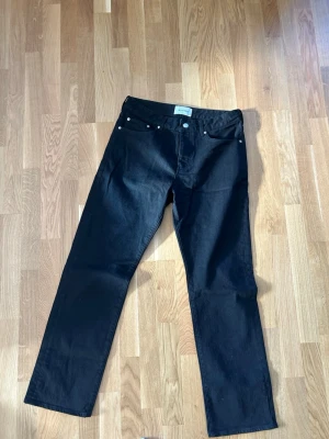 Svarta raka jeans 33x32 whyred  - Säljer ett par svarta jeans med rak passform och klassisk femficksdesign. Jeansen har silverfärgade nitar och knapp, samt bälteshällor i midjan. Materialet är kraftigt denim som ger en stilren look. Straight fit whyred helt nya aldrig använda 