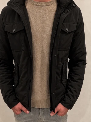J.Lindeberg Svart Field Jacka - Svart J.Lindeberg Field Jacket i storlek M. Jackan är vadderad, har två bröstfickor samt två sidofickor. Observera att jackan säljs med tydliga defekter: två-tre knappar framtill saknas och dragkedjan fungerar inte, vilket innebär att jackan inte går att stänga framtill i nuvarande skick. Säljs därför som defekt och till ett lägre pris. I övrigt ser jag inga andra större fel. Se bilderna noggrant före köp och skriv gärna vid frågor.