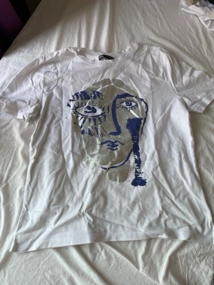 T-shirt med tryck - Säljer en vit t-shirt med ett stort ansiktstryck i blått och grått på framsidan. T-shirten har rund hals och korta ärmar. Pris kan diskuteras! Köpt från en annan säljare, men den var för stor.