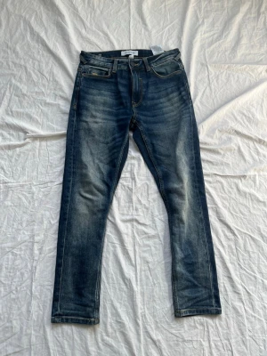 Mörkblå jeans från SMOG/DENIM - Snygga mörkblå jeans från SMOG/DENIM med klassisk femficksmodell och slitna detaljer på låren. Jeansen har smal passform och är tillverkade i denim med kontrastsömmar. Perfekta för en trendig och avslappnad stil.